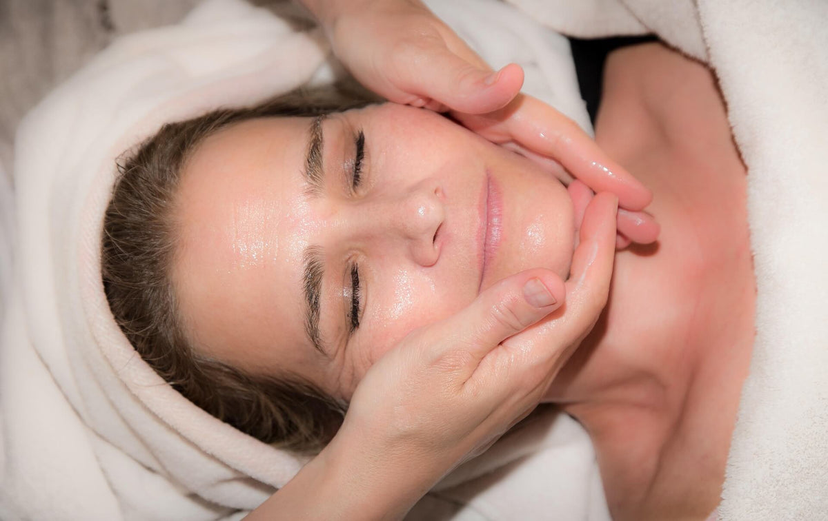 A close-up of a woman receiving a facial massage, wrapped in a white towel with her eyes closed as hands gently cleanse her face.